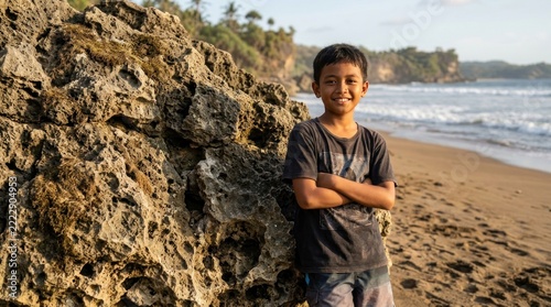 Smiling indonesian boy poses arms crossed by large beach rock