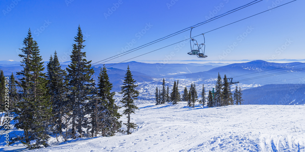 Naklejka premium Winter panoramic view of Sheregesh ski resort in Russia. Cable car ascends above the fir conifer forest and mountain valley, with houses hotels and distant peaks below, banner scenery, Green mount.