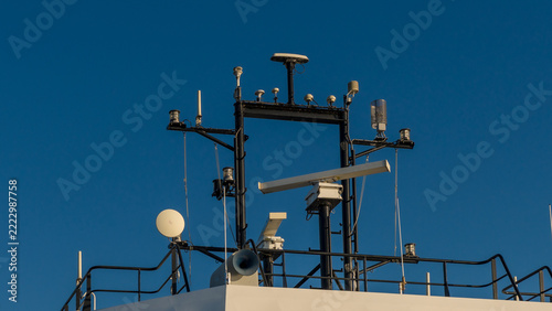 Ferry upper deck with railings, antennas, satellite dish, loudspeaker, and navigation equipment against a clear blue sky at sea