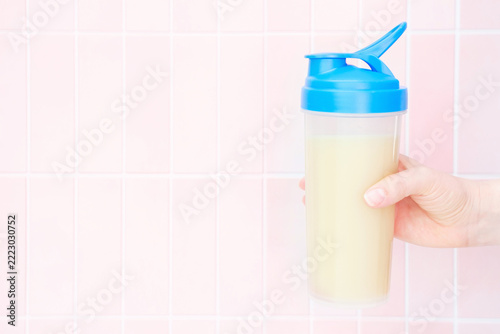 Female hand holds shaker with protein shake against background of pink tiles.