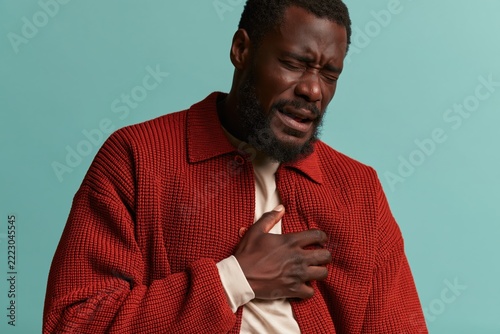Man holding his chest with a pained expression, experiencing heart pain or discomfort in a studio setting with a blue background