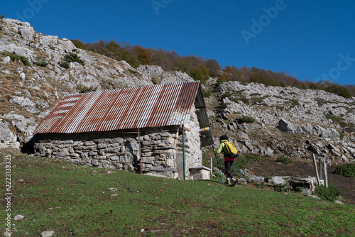 a hiker with backpack and trekking poles walks along a grassy trail beside a traditional old stone shelter with a rusty corrugated metal roof set against rocky mountain slopes under a clear blue sky.