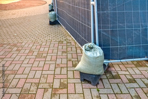 • Temporary construction fence with green mesh secured with heavy sandbags on sidewalk brick pavement. City infrastructure backdrop.