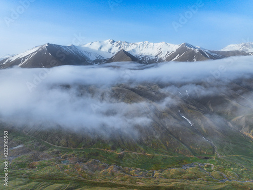 Thick layer of fog and clouds hovering across grassy hills and snowy summits