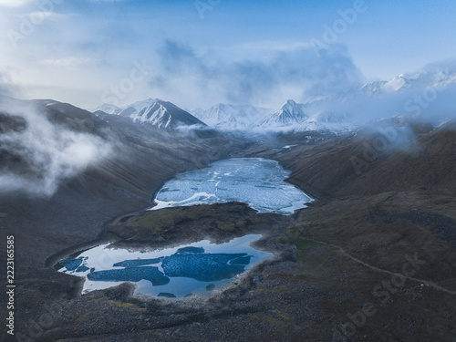 High angle view of mountain lakes with patches of ice in a rocky valley beneath low clouds and snowy peaks