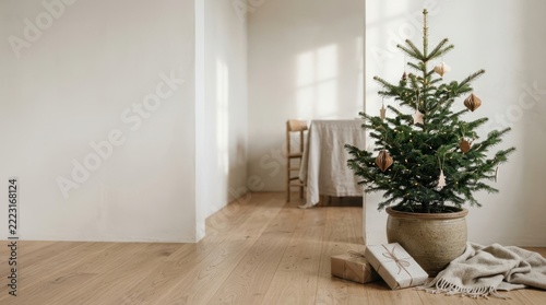 Minimalist interior scene featuring a small potted Christmas tree adorned with delicate paper ornaments and string lights, accompanied by wrapped gifts and a cozy blanket on wooden flooring