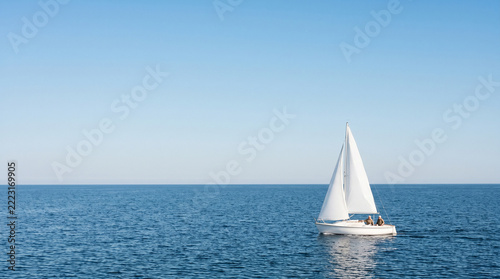 An elderly couple sails a small white sailboat across the blue sea under a cloudless blue sky; the older man and woman are enjoying a boat trip and an active retirement—a minimalist seascape