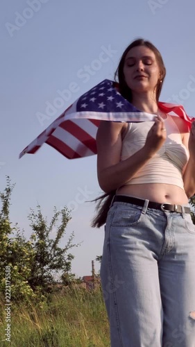 Young woman holding waving US flag on her shoulders outdoors in field. Concept of Independence Day, 4th of July, American patriotism, freedom, national holiday, democracy and USA pride