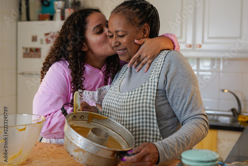 Happy african daughter kissing mother inside kitchen while preparing cake - Family people, bonding and food concept - Soft focus on girl hand - UGC content