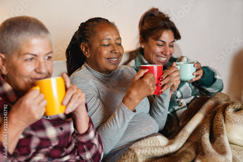 Happy multiracial senior women drinking hot tea together while sitting on couch during winter time - Cozy, elderly women friendship and lifestyle concept
