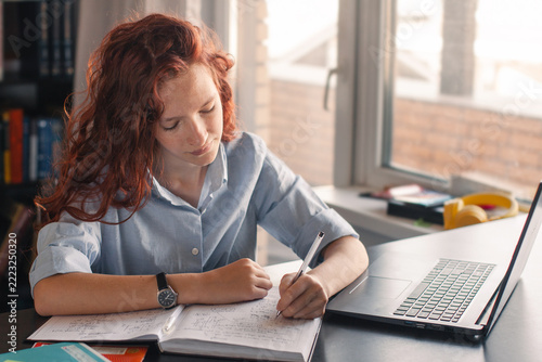 Teen girl writing notes in an open notebook beside a laptop at a desk. Student studying and completing schoolwork. Education, homework and digital learning concept. Academic focus.