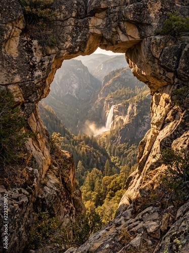 Breathtaking view of a cascading waterfall in a deep forested valley framed by a rugged natural stone archway under warm golden sunlight