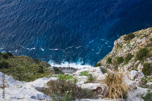  Dramatic view from a rocky cliff descending toward the deep blue Mediterranean Sea. Natural coastal landscape featuring limestone rocks, rugged vegetation and waves breaking along the shoreline.
