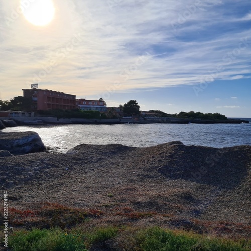  Coastal landscape illuminated by warm sunlight reflecting on calm waters. Waterfront buildings, shoreline and expansive sky create a relaxing Mediterranean atmosphere.
