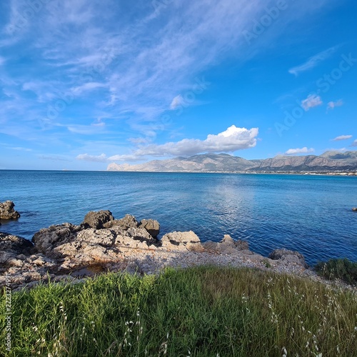  Wide coastal panorama featuring rocky shores, green vegetation, clear sea and impressive mountain scenery beneath bright blue skies. Perfect representation of southern European nature.

