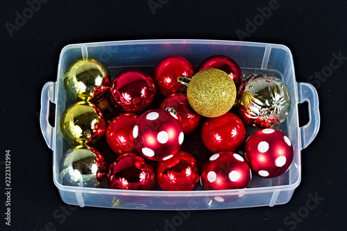 Glossy red ornaments, gold glitter spheres, and patterned baubles fill a transparent handled container, with bright reflections against a dark backdrop.