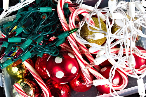 Red ornaments with white dots, striped candy canes, gold glitter and green and white Christmas lights fill a crowded holiday decoration box.