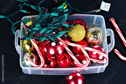 Red, gold, and silver ornaments fill a clear storage container, while tangled Christmas lights and striped candy canes spill onto a dark surface.