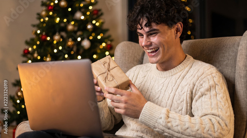 Surprised gift recipient laughing while unwrapping online order in armchair by Christmas tree