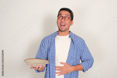 Asian man holding empty dining plate and touching his stomach with excited expression