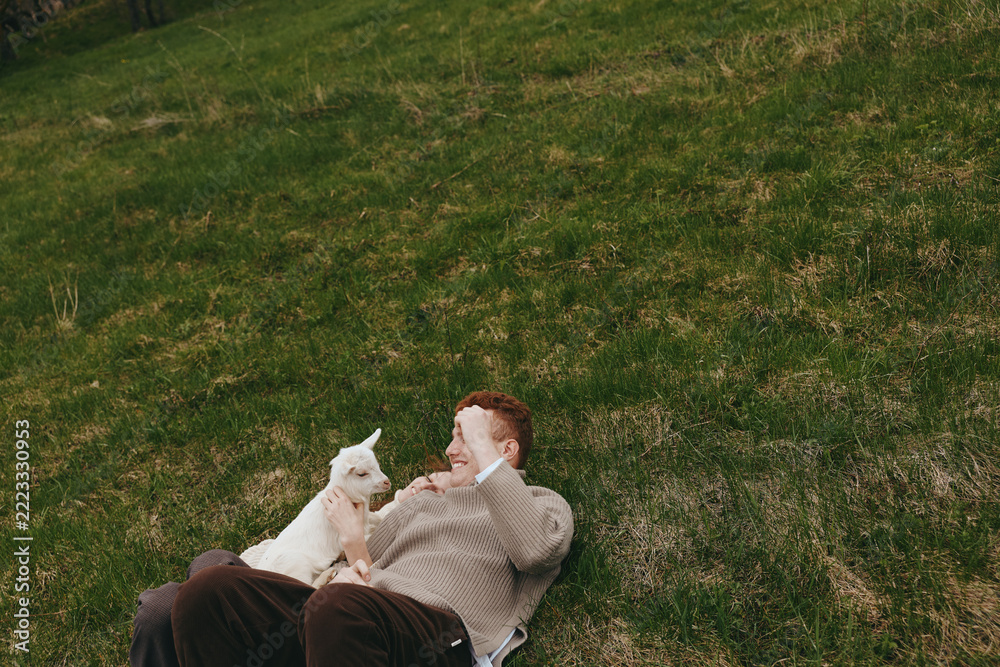 Fototapeta premium Cheerful man relaxing outdoors with a curious goat kid beside him, sunlit grass, casual clothing, warm smile, peaceful moment, nature setting, candid lifestyle scene