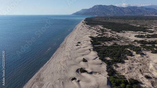 Aerial motion of expansive coastal sand dunes rolling parallel to calm blue sea with scattered green scrub and distant mountain range under clear sky, camera gliding along shoreline to reveal wind-swe