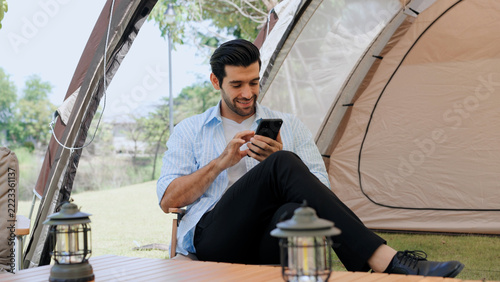 Happy young man using smartphone while relaxing outside his camping tent in nature, enjoying mobile connectivity, digital nomad lifestyle, outdoor recreation, and peaceful vacation time
