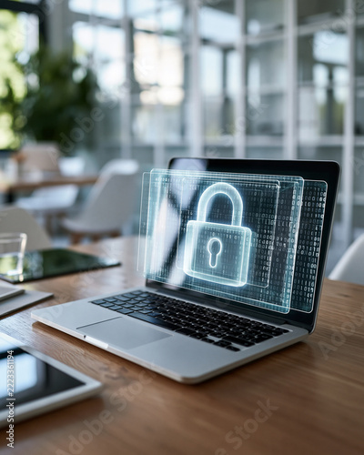 Modern office space with sleek laptop. Laptop screen displays glowing digital padlock. Futuristic interface suggests cybersecurity or data protection. Natural light streams through large glass windows