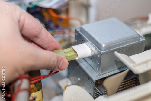 Hand connects a yellow plug to a microwave magnetron inside an open appliance. Silver metal housing and wiring are visible during repair