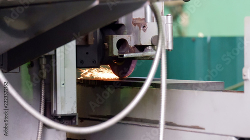 An industrial metal grinding machine in operation, with a rotating grinding wheel producing sparks while grinding a metal workpiece inside a manufacturing factory.