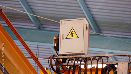 An industrial electrical control cabinet with a high voltage warning symbol, mounted on a steel structure inside a manufacturing factory.