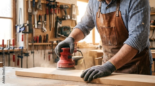 Carpenter using electric sander on wooden board in workshop with woodworker woodworking carpentry craftsman