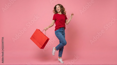 Joyful young woman in red t-shirt and jeans carrying a large red shopping bag against a vibrant pink background