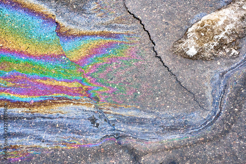Multicolored oil residue streaks across asphalt near stained rock. Cracked pavement holds vivid iridescent layers and dark speckles, showing abstract surface pattern associated with pollution.