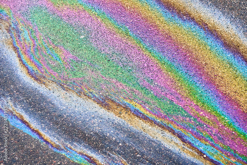 Iridescent petroleum film spreads across coarse asphalt surface, shimmering with vivid spectra. Liquid rainbow reflection highlights urban road pollution across textured ground texture