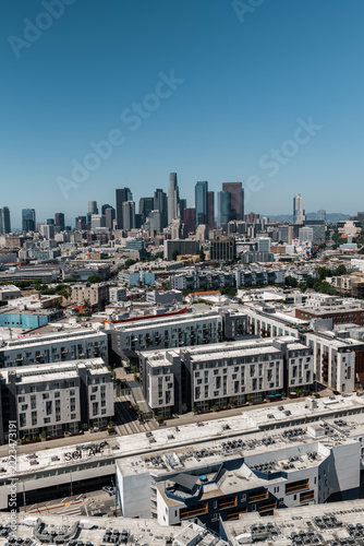 Aerial perspective reveals the urban sprawl and modern residential complexes of the Arts District leading toward the downtown Los Angeles skyscraper skyline under a clear afternoon sky.