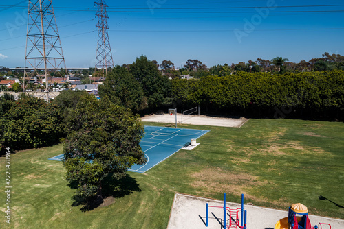Aerial shot reveals a public community park complete with outdoor basketball court, volleyball sand pit, lawn, children playground equipment, and electrical power towers.