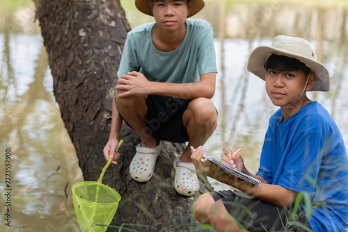 Two Asian high school students conducting a biology field research by a pond, with one boy writing observation notes on a notebook while his friend holds a scoop net.