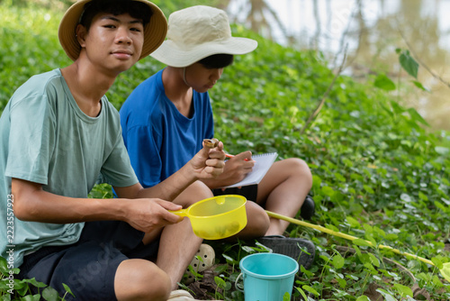 An Asian teenage student holding and examining a small aquatic specimen or shell found in a pond, sitting on green grass with his friend taking notes during a field trip.