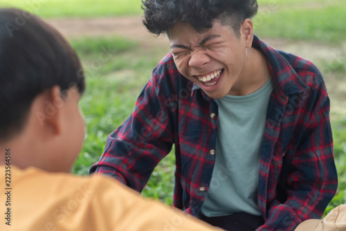 Close-up shot of a happy Asian teenage boy laughing out loud while chatting with his friend on green grass outdoors, concept of pure joy, youth friendship and positive emotion.