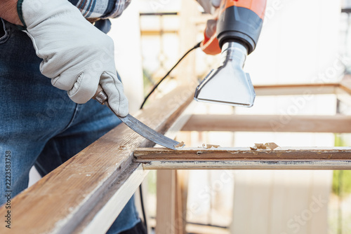 Adult carpenter wearing protective leather gloves, using a heat gun and a wood scraper to remove paint from an old wooden window. Construction industry, carpentry. Restoration. 