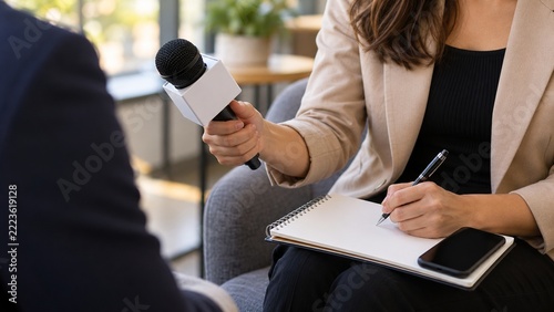 Female journalist holds a microphone and writes notes in a notebook while interviewing a business professional in a modern office lounge, capturing information for a media reporting assignment.