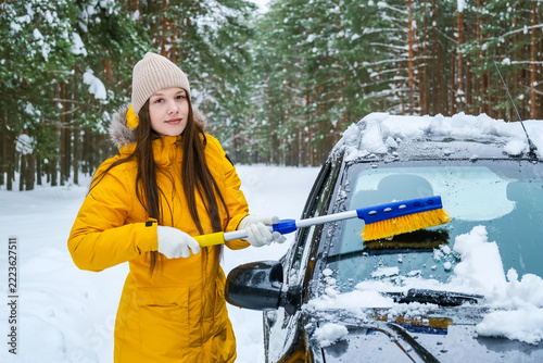 Young woman clearing snow from car with brush in winter forest. Snow removal, vehicle maintenance and safe driving preparation during cold weather concept.
