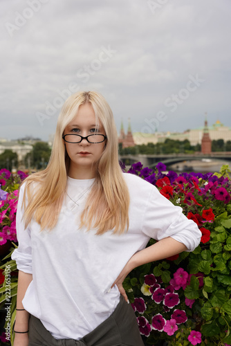 Young blonde woman with glasses wearing funny cat print sweatshirt standing outdoors among colorful flowers. Urban lifestyle and humorous street fashion concept with city background