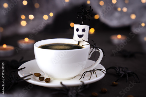White cup of coffee decorated with playful Halloween spiders and a smiling marshmallow ghost. Dark background, glowing candles and spiders in the background. Close-up.