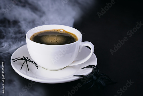 White cup of black coffee with decorative black spiders on a saucer. Dark background and mysterious smoke creating a spooky Halloween atmosphere. Close-up. Copy space.