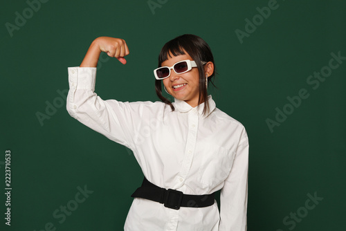 Smiling Teenage Girl Wearing Sunglasses and Flexing Biceps Against Green Background