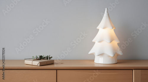 Serene minimalist scene with a white ceramic Christmas tree on a light oak table. Simple gift with eucalyptus. Clean, Scandinavian, and peacefully modern holiday decor.
