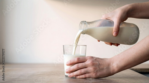Hands pour fresh white milk from a glass bottle into a clear glass, a simple healthy morning routine with basic food on a clean neutral background.