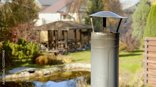 Metal chimney pipe stands on roof overlooking countryside backyard. Close up focuses on steel cowl with blurred garden pond, gazebo, and cottage in sunny autumn afternoon.